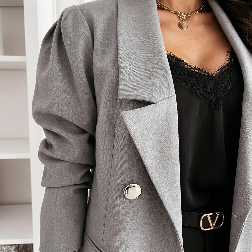 A close-up of a person wearing a light gray double-breasted blazer with dramatic puffed sleeves, layered over a black lace-trim camisole and a black belt with a gold-tone V-shaped buckle.