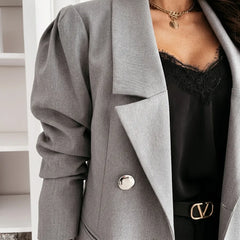 A close-up of a person wearing a light gray double-breasted blazer with dramatic puffed sleeves, layered over a black lace-trim camisole and a black belt with a gold-tone V-shaped buckle.