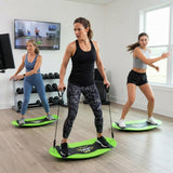 Three women exercise indoors on bright green and black board-walking devices in a fitness studio with wood floors, dumbbells, and a window.