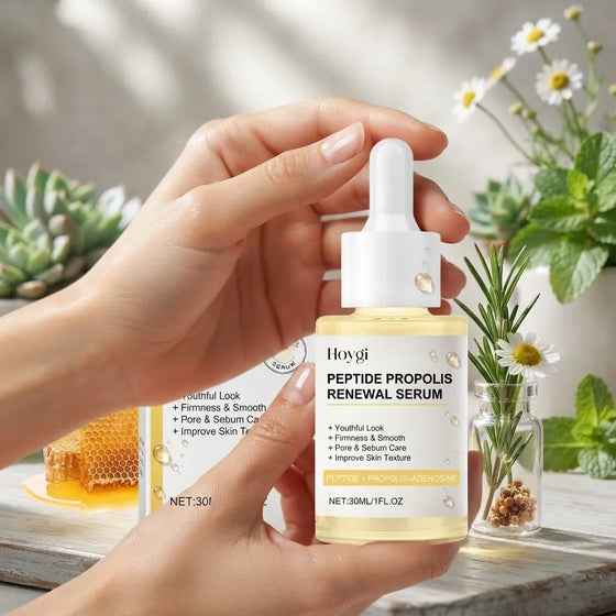 A person's hands hold a bottle of Hoygi Peptide Propolis Renewal Serum, which is a pale yellow liquid in a clear bottle with a white dropper cap. The background is a beautifully styled, soft-focus scene featuring a glass jar of honey, fresh daisies in a small vase, and green succulents on a rustic wooden table.