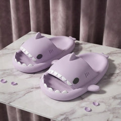 A pair of lavender purple shark-themed slide slippers displayed on a white marble-patterned surface with a soft mauve curtain background.