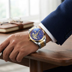 A luxury men's watch with a deep blue dial and gold-tone case, worn on a man's wrist. The watch features a two-tone silver and gold stainless steel link bracelet.