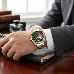 A man wearing a two-tone gold and silver wristwatch with a black dial, resting his hand on a dark leather notebook at a wooden desk.