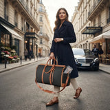 A woman in a dark navy coat walks down a city street carrying a dark gray duffel bag with tan leather trim and straps. A luxury hotel sign and a black car are visible in the background.