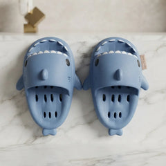 A pair of light blue shark-shaped clogs with white teeth details, displayed on a white marble surface.