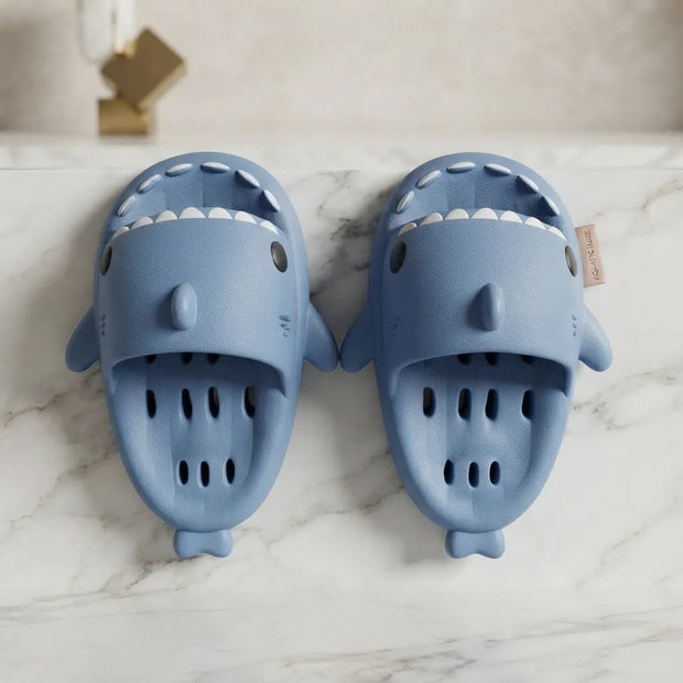A pair of light blue shark-shaped clogs with white teeth details, displayed on a white marble surface.