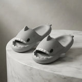 A pair of gray shark-shaped slide sandals displayed on a white marble pedestal against a neutral gray background.