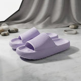 A pair of light purple textured slide slippers on a white marble surface with a draped gray fabric backdrop and small decorative stones.