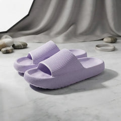 A pair of light purple textured slide slippers on a white marble surface with a draped gray fabric backdrop and small decorative stones.