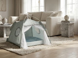 A small dome pet play tent in a pale sage green and white patterned fabric with sheer white tulle curtains, set up on a beige shag rug in a bright, modern living room with a cream sofa and marble side tables.