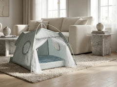 A small dome pet play tent in a pale sage green and white patterned fabric with sheer white tulle curtains, set up on a beige shag rug in a bright, modern living room with a cream sofa and marble side tables.