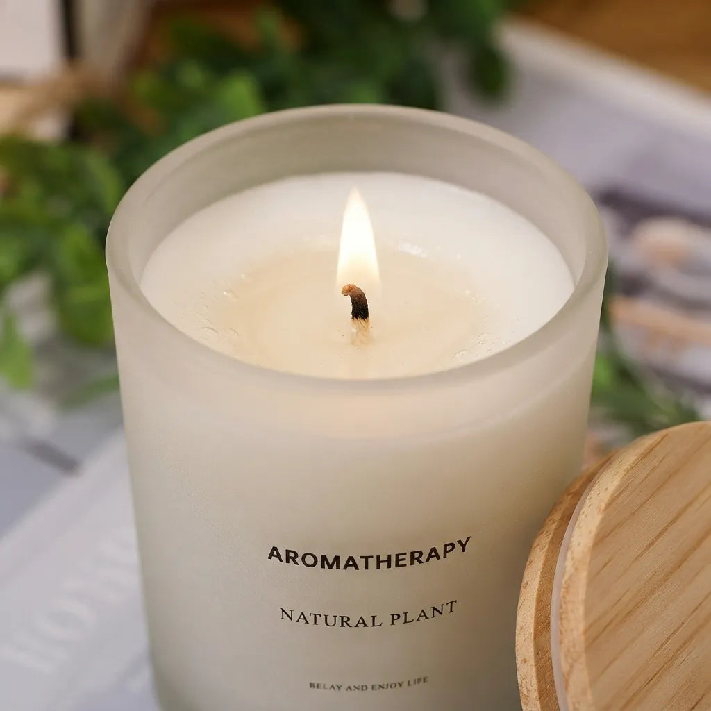 A frosted glass candle with a lit wick, labeled 'AROMATHERAPY NATURAL PLANT,' shown in a softly blurred setting with a wooden lid partially visible at the right.