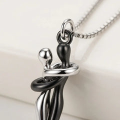 A close-up of a pendant necklace featuring two intertwined human figures in polished silver and matte black finishes, suspended from a silver chain against a soft off-white background.