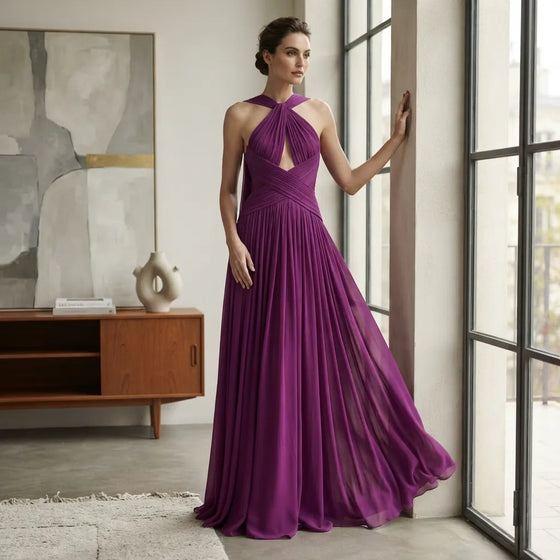 A woman stands indoors by large windows wearing a flowing purple halter-neck maxi dress with a keyhole cutout at the chest and a pleated skirt. The room has a wood cabinet, abstract wall art, and a light rug.