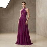 A woman modeling a floor-length plum purple halter gown with a keyhole cutout at the chest, standing in a minimalist studio with textured beige walls.