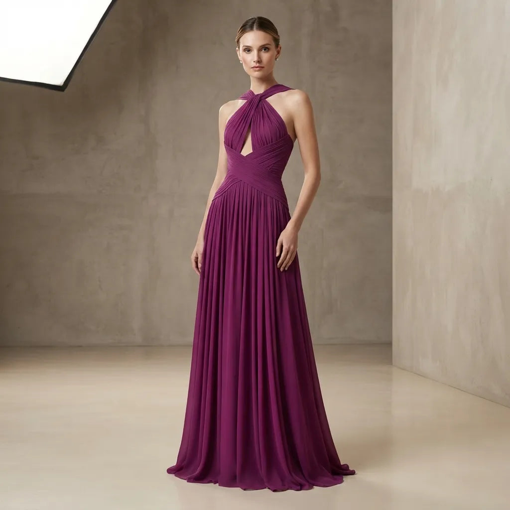 A woman modeling a floor-length plum purple halter gown with a keyhole cutout at the chest, standing in a minimalist studio with textured beige walls.