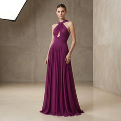 A woman modeling a floor-length plum purple halter gown with a keyhole cutout at the chest, standing in a minimalist studio with textured beige walls.