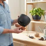 A person in a light blue denim shirt holds a matte black hanging planter pot with a brown saucer and chain, standing in a bright garden center or home garden setting with a wooden table, soil, twine, and another potted plant on a shelf in the background.