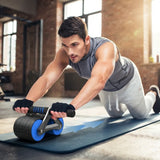 A man in a grey sleeveless shirt and white sweatpants performs an abdominal exercise on a blue exercise mat using a black and blue ab roller wheel in a gym with brick walls.