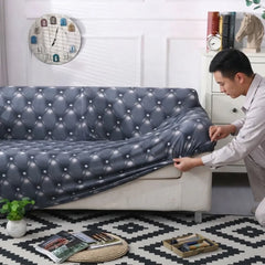 A man in a white shirt is shown pulling a dark gray tufted sofa slipcover onto a light beige sofa in a decorated living room setting.
