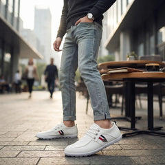 A person standing outdoors in a city setting wearing light-wash blue jeans and white low-top sneakers with a small red, white, and blue stripe detail on the side. The background shows blurred tables, chairs, and pedestrians.