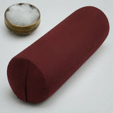 A cylindrical red fabric yoga bolster pillow shown on a light gray background, with a small brass bowl containing white cotton padding in the upper left corner.