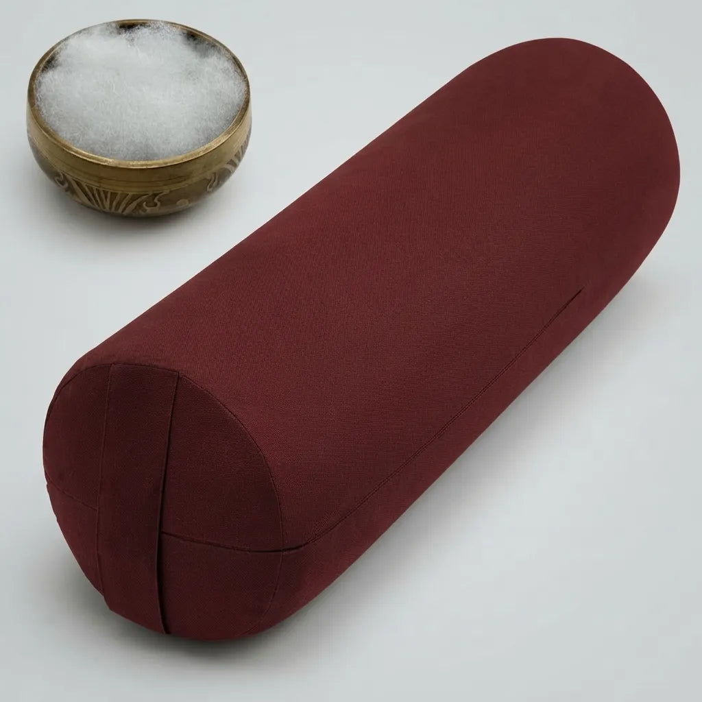 A cylindrical red fabric yoga bolster pillow shown on a light gray background, with a small brass bowl containing white cotton padding in the upper left corner.