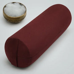 A cylindrical red fabric yoga bolster pillow shown on a light gray background, with a small brass bowl containing white cotton padding in the upper left corner.