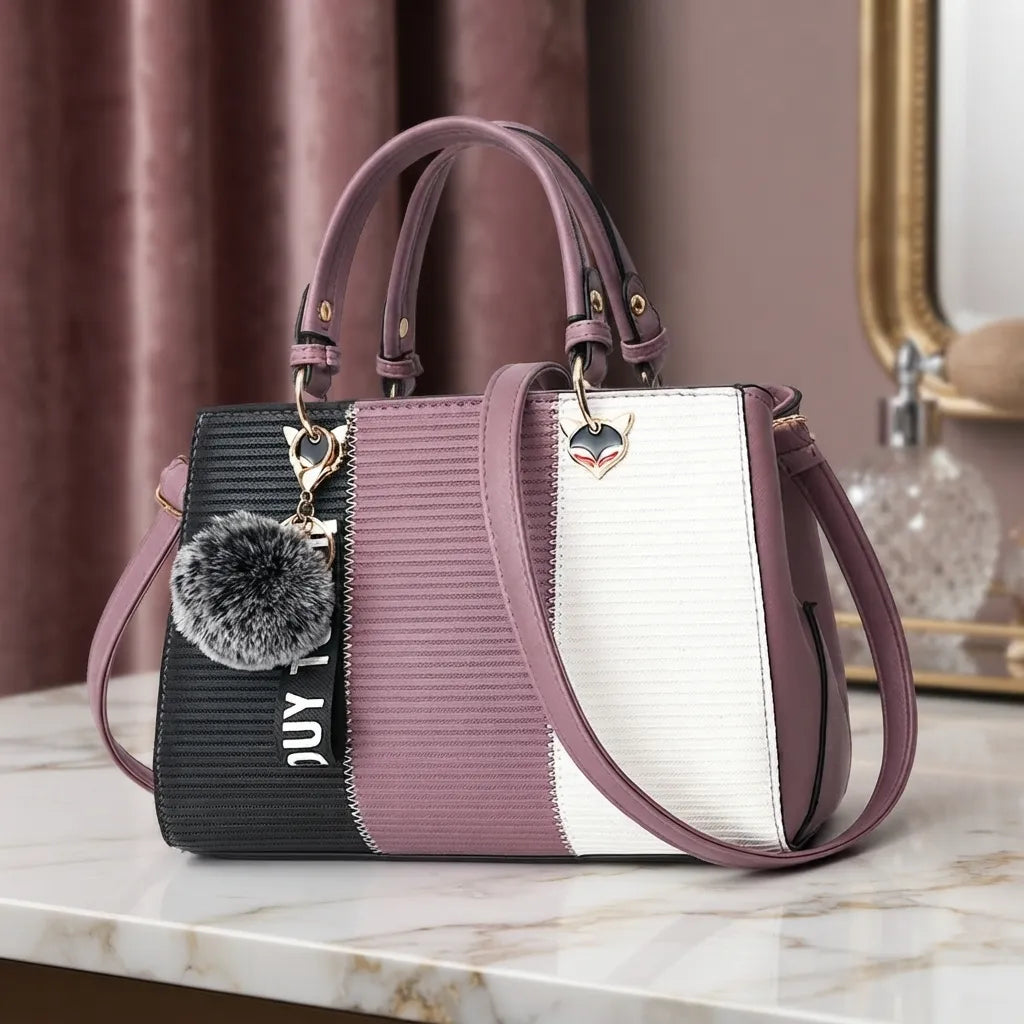 A structured handbag with a color-blocked design in black, textured mauve, and white, featuring dual top handles, a shoulder strap, gold-tone hardware, and a gray faux-fur keychain charm.