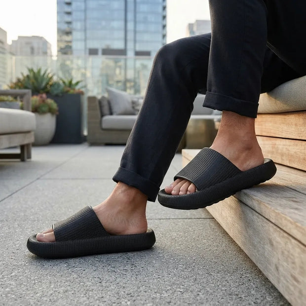 A person sitting on outdoor wooden steps wears black slide sandals with wide ribbed straps and thick cushioned soles, with dark pants and a modern rooftop patio in the background.