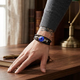 A close-up of a person's hand resting on a wooden desk, wearing a multi-strand black leather bracelet with a central round blue stone and metallic spacer beads.