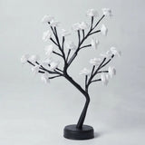 A decorative artificial flowering tree with black branches and white blossoms on a round black base, photographed against a plain light gray studio background.