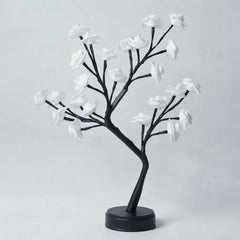 A decorative artificial flowering tree with black branches and white blossoms on a round black base, photographed against a plain light gray studio background.