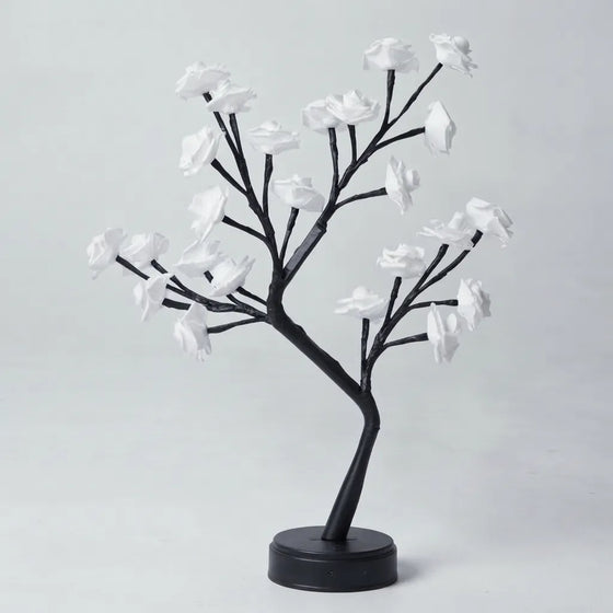 A decorative artificial flowering tree with black branches and white blossoms on a round black base, photographed against a plain light gray studio background.