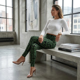A woman sits on a concrete bench in a modern loft setting, wearing high-waisted green and black geometric patterned leggings with a white ribbed long-sleeve top and clear high-heel mules.