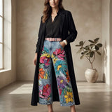 A woman stands in a sunlit room wearing a long black coat over a black blouse, paired with wide-leg denim jeans covered in large colorful floral prints and a pink belt. A potted plant is visible in the background.