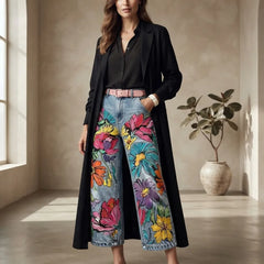 A woman stands in a sunlit room wearing a long black coat over a black blouse, paired with wide-leg denim jeans covered in large colorful floral prints and a pink belt. A potted plant is visible in the background.