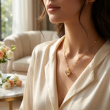 A close-up lifestyle shot of a woman wearing a delicate gold heart pendant necklace with a central crystal, styled with a cream satin blouse in a softly lit, elegant room.