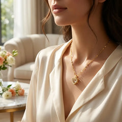 A close-up lifestyle shot of a woman wearing a delicate gold heart pendant necklace with a central crystal, styled with a cream satin blouse in a softly lit, elegant room.
