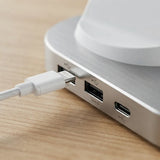 Close-up of a white braided USB-C cable plugged into the side ports of a brushed silver aluminum hub, resting on a light wood surface.