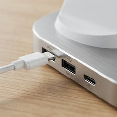 Close-up of a white braided USB-C cable plugged into the side ports of a brushed silver aluminum hub, resting on a light wood surface.