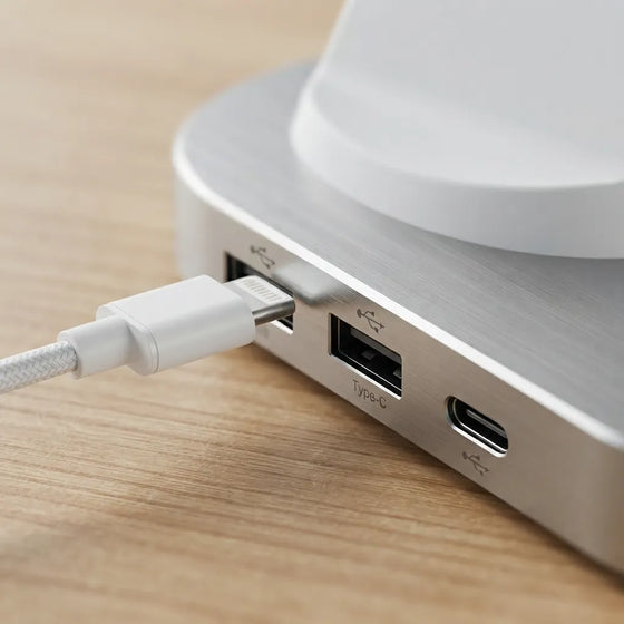 Close-up of a white braided USB-C cable plugged into the side ports of a brushed silver aluminum hub, resting on a light wood surface.