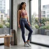A woman stands indoors by large windows wearing a dusty rose sports bra and dark navy patterned leggings with white sneakers. The setting includes a concrete pillar, a small wooden table with a water bottle, and a rolled mat.