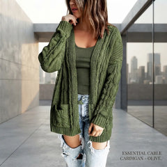 A woman wearing an olive green cable-knit cardigan over a matching green top and distressed light-wash jeans, standing in a modern concrete walkway with a city skyline visible through glass.