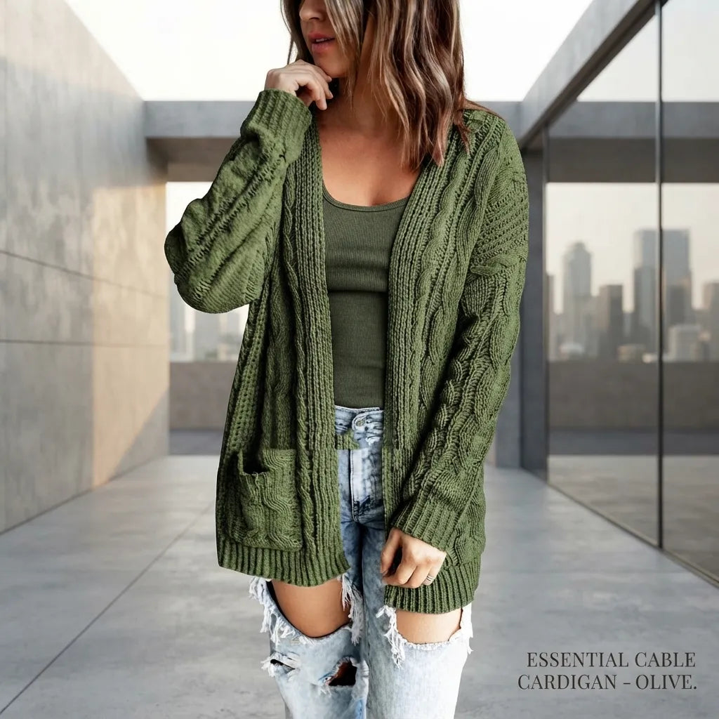 A woman wearing an olive green cable-knit cardigan over a matching green top and distressed light-wash jeans, standing in a modern concrete walkway with a city skyline visible through glass.