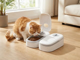A white dual pet feeder and waterer with an open lid sits on a light wood floor next to an orange and white cat eating dry food, in a bright living room setting.