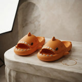 A pair of orange shark-shaped slide slippers with white teeth details, displayed on a light marble table next to a delicate gold chain.