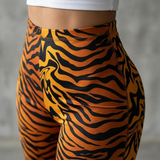 Close-up of a person wearing high-waisted tiger-print leggings in orange and black, paired with a white ribbed waistband top, against a soft gray concrete background.