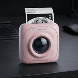 A pink SQUARE paperless printer sits on a dark desk with a printed sheet emerging from the top. The device has a glossy black circular lens area, a power button, and the brand name PAPERANG printed above it.