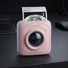 A pink SQUARE paperless printer sits on a dark desk with a printed sheet emerging from the top. The device has a glossy black circular lens area, a power button, and the brand name PAPERANG printed above it.